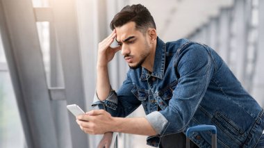 Endişeli genç Arap adam havaalanı terminalinde beklerken akıllı telefon ekranına bakıyor, hayal kırıklığına uğramış Orta Doğulu adam alnına dokunuyor, kötü mesaj okuyor ya da internet bağlantısı sorunu yaşıyor.