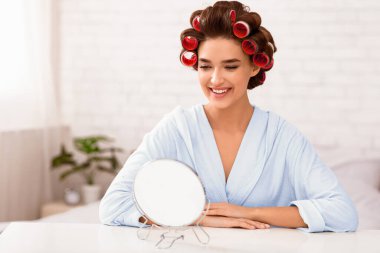 Saç şekillendirme. Evdeki modern yatak odasında, yuvarlak aynanın önünde oturan kıvırcık saçlı çekici kadın. Kadın Güzellik Rutini Konsepti. Metin için Seçici Odaklanma, Boşluk Boşluğu