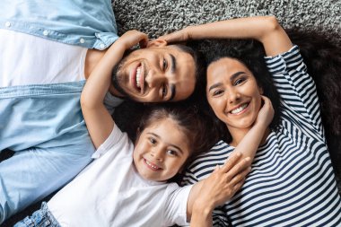 Evlerinde yerde poz veren mutlu orta doğu ailesinin yakın plan portresi, halının üzerine uzanmış, birbirine bağlanmış, neşeyle gülümseyen, yukarıdan bakan. Pozitif baba, anne-kız kucaklaşması, üst bakış.