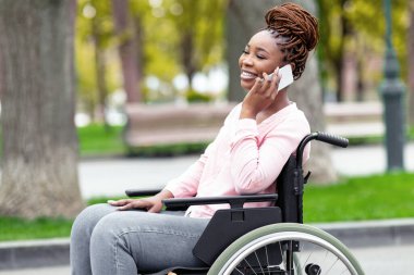 Tekerlekli sandalyedeki güzel siyahi bayan sonbaharda şehir parkında akıllı telefondan konuşuyor. Belden aşağısı felçli Afro bir kadın sıcak bir sonbahar gününde dışarıda cep telefonuyla konuşuyor.