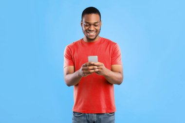 Mavi Stüdyo Arkaplanının Üzerinde Duran Cep Telefonu Mesajı Kullanarak Afro-Amerikalı Mutlu Erkek. Neşeli siyahi adam internette geziniyor. Akıllı telefonunuz için yeni bir uygulama. İnsanlar ve Gadget
