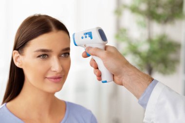 Yeni Normal, Sıcaklık Denetimi. Temassız Infrared Themometer Silah Ölçme Ateşi, Klinikteki Genç Kadının Alnı Taranıyor. Coronavirus, Covid-19 Virüs Yayılma Kontrolü
