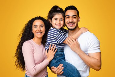 Küçük kızıyla birlikte mutlu bir orta doğu ailesi kameraya sarılıp gülümsüyor, Arap ebeveynleri seviyor ve sarı stüdyo sırt çantasının üzerinde tatlı bir çocuk pozu veriyorlar.