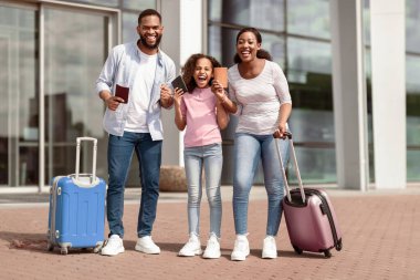 Sonunda tatil zamanı. Biletleri olan ve yumruklarını sıkmış üç kişilik neşeli, pozitif siyah aile, kazanan jest yapıyor, havaalanı terminalinin yanında birlikte duruyorlar.