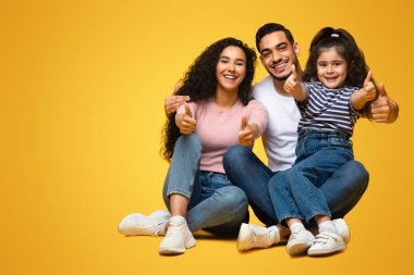 Tavsiye ediyoruz. Kamerada üç çocuklu mutlu bir Arap ailenin portresi, neşeli bir Orta Doğulu ebeveyn ve küçük bir kızın onaylama işareti göstermesi, sarı arka planda poz vermesi, boşluğu kopyalaması.