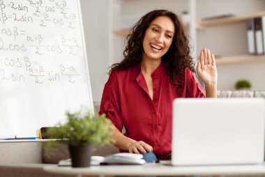 Uzaklık Eğitimi. Pozitif kadın portresi sanal toplantı yapıyor, masada oturuyor, öğrencilerle bilgisayar başında video görüşmesi yapıyor, webcam 'e el sallıyor. Evde matematik dersi veren bir kadın.