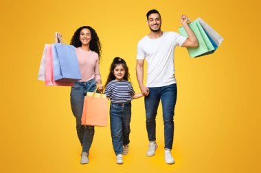 Neşeli Arap ebeveynler ve parlak alışveriş torbaları olan küçük kızları sarı stüdyo arka planında yürüyen, üç satın alma ve gülümseme taşıyan neşeli ortadoğulu aile,