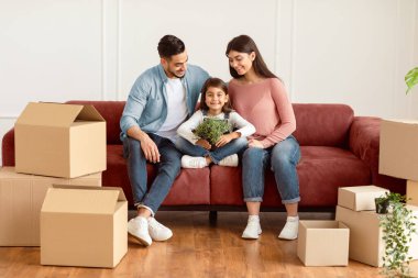 Yeni Apartmanda. Mutlu Ortadoğulu aile taşınma gününü kutluyor, elinde bitki tutan heyecanlı kız, koltukta oturan ebeveynler ve yerde karton kutular olan kızlarına bakıyor.