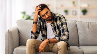 Stresli sakallı Arap adam gözlüklü, gözlüklü, evde tek başına oturuyor, kafasına dokunuyor ve aşağı bakıyor, akşamdan kalma ya da zorluk yaşıyor, boşluğu kopyalıyor.