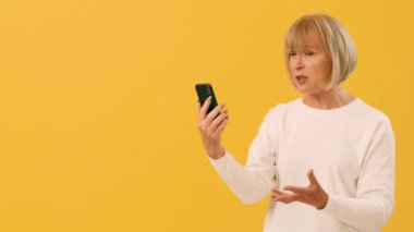 Kısa saçlı bir kadın, canlı bir şekilde akıllı telefonuna konuşuyor. Parlak sarı bir arkaplan önünde dururken duygularını gösteriyor. Çağrı sırasında meşgul ve anlamlı görünüyor..