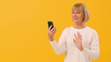 Yaşlı bir kadın akıllı telefonuyla yaptığı bir video görüşmesinin keyfini çıkarırken gülümser ve el sallar, canlı sarı bir arka plana kurulur, iletişiminde neşe ve bağlantı gösterir, boşluğu taklit eder.
