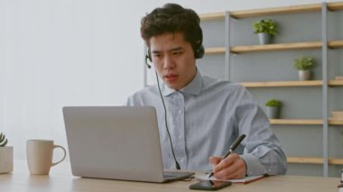 Genç bir Asyalı adam ev ofisinde uzaktan çalışmakla meşgul. Dizüstü bilgisayarında yazarken kulaklık takıyor ve görevlerine odaklanmış bir not defterine notlar yazıyor..