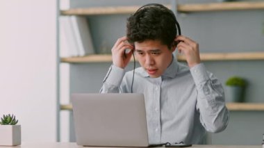 Yakalı gömlek giymiş genç bir Asyalı adam, çağdaş bir merkez ofisteki bir masada oturuyor. Kulaklık tutarken dikkatle dizüstü bilgisayarına bakıyor..
