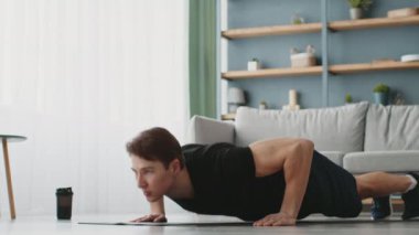 Genç, sportif bir adam şık oturma odasındaki paspasın üstünde şınav çekiyor. Antrenmanına odaklanırken doğal ışık boşluğu dolduruyor. Yakınlarda bir kahve fincanı duruyor, rahatlatıcı atmosferi arttırıyor..