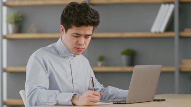 Genç bir Asyalı adam sıcak bir ofiste uzaktan çalışmakla meşgul. Dizüstü bilgisayarına odaklanıyor, bitkiler ve kitaplarla çevrili bir şekilde not alıyor..