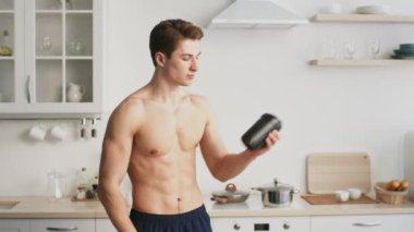 Formda bir genç adam şık mutfağında durmuş protein içeceği kabını inceliyor. Sağlıklı bir yaşam tarzını ve bir anlık yansımanın tadını çıkarırken spor yapmaya bağlılığını somutlaştırıyor..