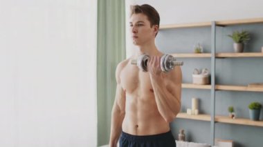 Genç, kaslı bir adam evde çalışıyor, odaklanmış bir dambılı kaldırıyor. Parlak çevre, spor yapmaya ve sağlıklı bir yaşam tarzına olan bağlılığı yansıtır..