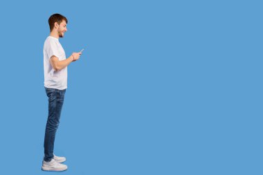 Cep telefonuyla mesajlaşan yakışıklı bir adamın tam boy yan görünüm portresi, Pastel mor stüdyo duvarı üzerinde izole edilmiş, uzay kopyası.