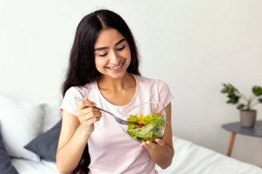 Neşeli Hintli kadın evde yatakta oturmuş taze sebze salatası yiyor. Güzel Hindu kadın vejetaryen yemeğinin tadını çıkarıyor. Temiz yaşam, sağlıklı beslenme, vegan beslenme konsepti