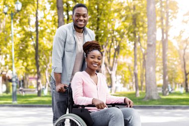 Sonbaharda tekerlekli sandalyedeki sakat kız arkadaşını şehir parkında yürüyüşe çıkaran neşeli siyahi bir adam. Afro-Amerikan engelli çift birlikte doğanın tadını çıkarıyorlar.