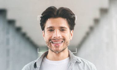 Gülümseyen genç bir adam, yumuşak ışıklandırmayla çevrili, modern bir koridorda duruyor. Yüzü dijital yüz tanıma teknolojisi tarafından çerçevelenmiş, tanımlama özellikleri vurgulanıyor.