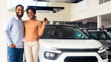 Showroom 'da lüks arabanın yanında poz veren mutlu Afrikalı Amerikalı eşler, neşeli siyah çift anahtarlarını tutuyor ve kameraya gülümsüyor, modern oto galerisinden yeni bir araç alıyor, tam boy, kopyalama boşluğu dolduruyor.
