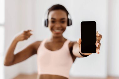 Fitness Uygulaması. Gülümseyen, spor kıyafetli genç Afrikalı Amerikalı kadın elinde cep telefonuyla izole edilmiş, boş ekranı işaret ederek şablon, seçici odak, bulanık arka plan.