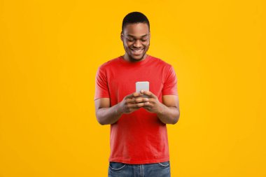 Gülümseyen Siyah Adam Smartphone Tarama İnternet ve Sosyal Medya Haber Kaynağı Sarı Stüdyo Arkaplanı 'nın üzerinde duruyor. Millennial Guy Yeni Mobil Uygulama Kullanıyor. İnsanlar ve Gadget