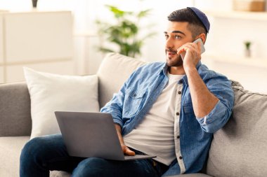 Telekomünikasyon ve Teleworking Concept. Genç Yahudi bir adamın portresi telefon görüşmesi yapıyor, bilgisayarda çalışıyor, oturma odasındaki kanepede oturuyor. Müşteri desteği. Dizüstü bilgisayar kullanan kendine güvenen yetişkin