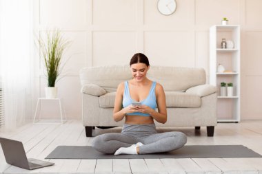 Cep telefonu egzersizi yapan genç bir kadın evdeki jimnastik minderinde oturuyor. Boşluğu Kopyala