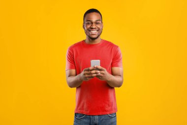 Yeni cep telefonu uygulamalı mutlu Afrikalı Amerikalı adam stüdyodaki sarı arka planda kameraya gülümsüyor. İnsanlar ve Gadget 'lar, Akıllı Telefon Konsepti için harika bir uygulama