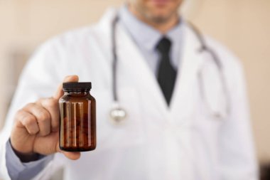 Doktor, parmaklarının arasında bir şişe hapla kameraya şeffaf kavanoz gösteriyor.
