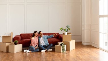 Gülümseyen bir ailenin portresi yerde oturuyor ve boş bir dairede kanepeye yaslanıyor. Boş alanı işaret eden adam karısını kucaklayan, duvar rengini ve iç tasarım tamirini seçen, pankart.