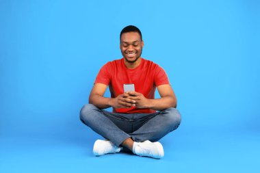 Mutlu Siyah Adam Cep Telefonu Kullanıyor İnterneti Tarıyor Mavi Arkaplanda Oturuyor. Stüdyo fotoğrafı, Millennial Guy 'ın Smartphone' da sosyal medya uygulamasını kullanması. Telefon kavramın için yeni uygulama