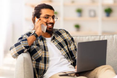 Pozitif uzun saçlı, sakallı, serbest çalışan bir Arap. Laptoyla koltukta oturuyor, telefon çekiyor, klavyede daktilo yazıyor, evden çalışıyor, ekrana bakıyor ve gülümsüyor, boşluğu kopyalıyor.