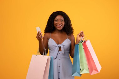 Joyful black woman using smartphone for online shopping, holding colorful shopper bags on orange studio background. Millennial African American female purchasing contactless via internet