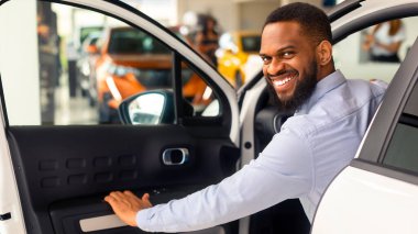 Gülümseyen Afrikalı Amerikalı adam yeni arabasının kapısını açıyor ve kameraya gülümsüyor, modern galeri merkezinde araba aldıktan sonra lüks bir araçta mutlu siyah erkek pozu veriyor,