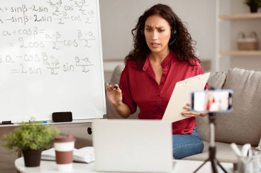 Çevrimiçi Çalışma, Uzaktan Öğrenme, Sanal Sınıf. Kablosuz kulaklık takan ciddi odaklı bayan öğretmen tripoddaki telefon kamerasıyla ilgili dersler ters çizelgenin yanında oturuyor, bilgisayar ekranı ile konuşuyor.