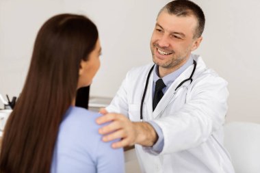 Doktor randevusundaki genç kadın. Mutlu erkek doktor hastasına iyi haberler veriyor, rahatlatıcı, elini omzunda tutuyor. Sağlık ve tıbbi konsept, yakın portre.