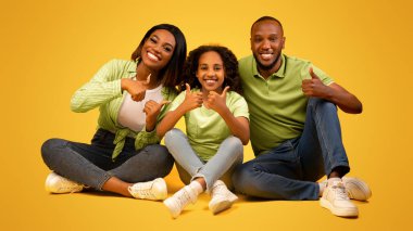 Üç kişilik Afro-Amerikan ailesi, sarı stüdyo arka planında oturarak bir şeyi onaylıyor ya da tavsiye ediyor.