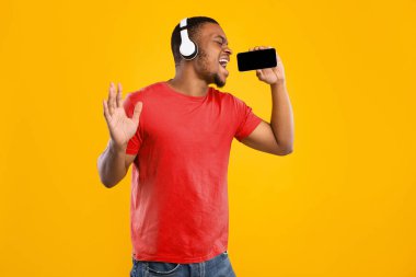 Siyah Adam Mikrofon Gibi Smartphone Söylüyor Karaoke Uygulamasını kullanarak Sarı Stüdyo Arkaplanında Dururken Eğleniyor. Kablosuz kulaklık takan Adam Müzik Uygulamasını Kullanarak Çevrimiçi Keyifli