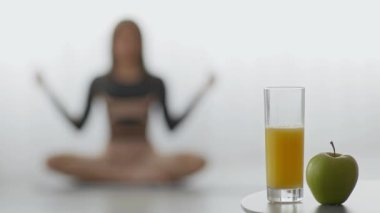 Bin yıllık Afro-Amerikan bir kadın ev ortamında yoga yapıyor. Formuna ve sağlığına odaklanıyor. Yakınlarda bir elma ve bir bardak meyve suyu var, sağlıklı bir yaşam tarzını destekliyor..