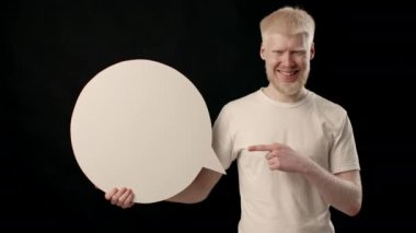 Albinizmi olan genç bir adam siyah bir geçmişi olan bir stüdyoda duruyor. Elinde tuttuğu büyük, boş bir konuşma balonunu işaret ederken gülümseyen beyaz bir teni ve saçı var..