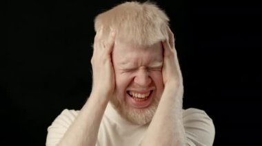 Albino özelliklere sahip genç bir adam, iki elini de koyu bir arka planda tutan, gözle görülür bir şekilde sıkıntılı. Beyaz teni ve parlak saçları etrafındaki karanlıkla tezat oluşturuyor..
