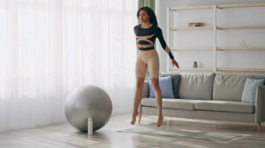 Aktif giyimli bir Afrikalı Amerikalı kadın oturma odasında çömelme gösterisi yapıyor. Doğal ışık perdelerin arasından akıyor, egzersiz sırasında kendini spora ve sağlığa adadığını vurguluyor..