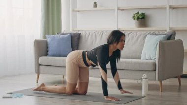 Genç bir Afrikalı Amerikalı kadın şık oturma odasında yoga minderi üzerinde egzersiz yapıyor. Rahat bir kanepe ve dekorla çevrili egzersiz rutinine odaklanıyor..