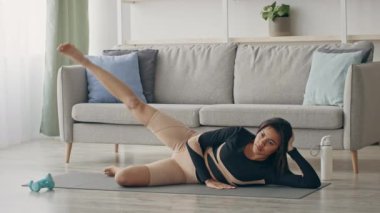 Bir kadın, iyi aydınlatılmış bir oturma odasında yan yatarak bacak kaldırma ameliyatı yapıyor. Spor giysileri içinde, bir halter ve halter kullanarak spor rutinine odaklanıyor, sağlığına olan bağlılığını gösteriyor..