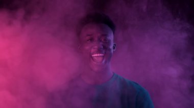 Genç bir Afrikalı Amerikalı şık bir görünümü olan adam canlı neon dumanların arasında kendinden emin bir şekilde gülümsüyor. Çarpıcı arka plan, onun modern estetik sergilenen havalı görünümünü arttırır..