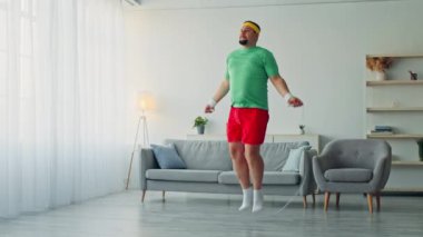 Tombul bir adam evde egzersiz yapıyor, parlak bir odada ip atlıyor. Yeşil gömlek ve kırmızı şort giyiyor, sağlıklı bir yaşam tarzını kucaklarken spor yolculuğuna odaklanıyor..