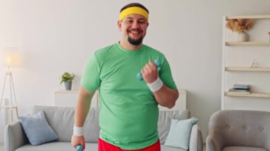Tombul bir genç adam aydınlık bir oturma odasında egzersiz yapıyor, halterleri kaldırırken gülümsüyor. Yeşil bir gömlek ve saç bandı takıyor, evdeki sağlık ve sağlık durumunu iyileştirmeye odaklanıyor..